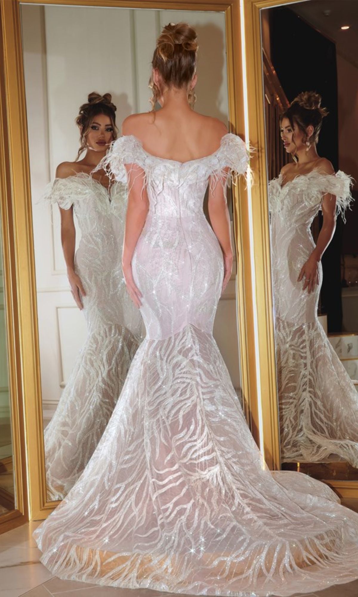 A woman in the Portia and Scarlett Long Prom Dress PS22227, an off-the-shoulder white mermaid gown with feather trim, stands before a large mirror, her elegant reflection visible from the front.